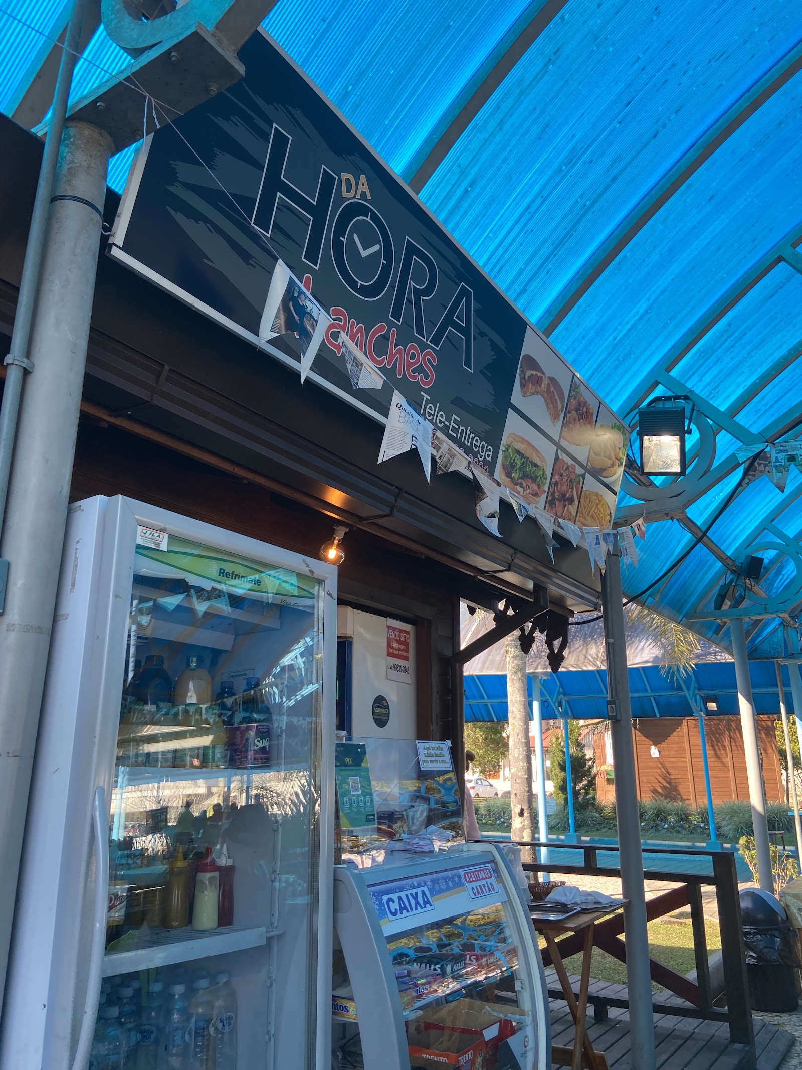 Placa e área de atendimento do Da Hora Lanches, sob cobertura azul · imagem de acervo de 2021.