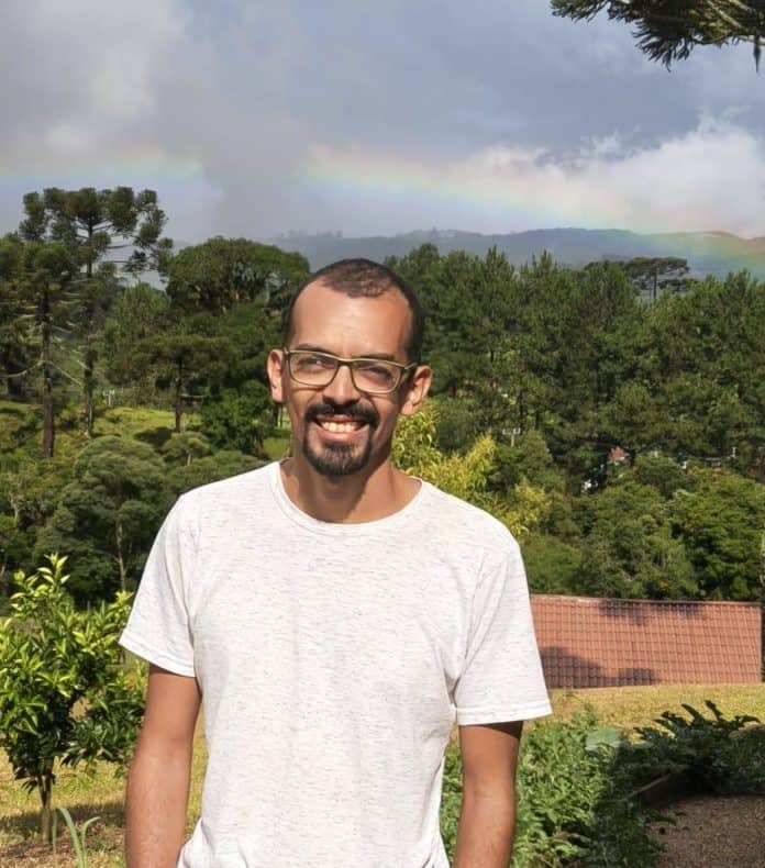 Felipe Novaes Rocha diante de araucárias e de um arco-íris