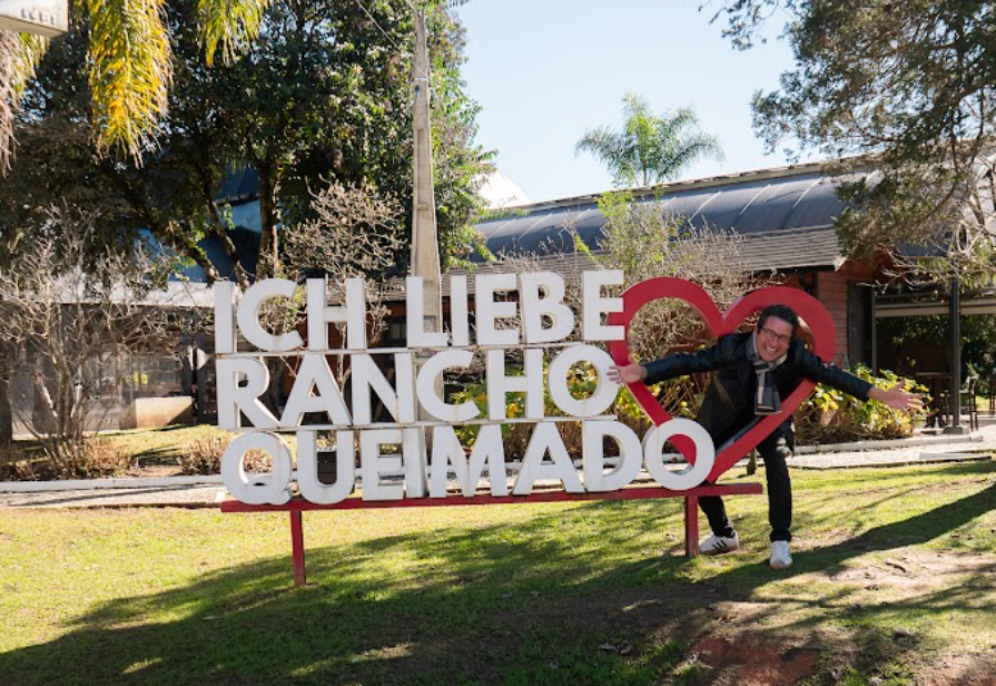 Capa da divulgação do vídeo da Modulare, com apresentador ao lado do letreiro Ich liebe Rancho Queimado
